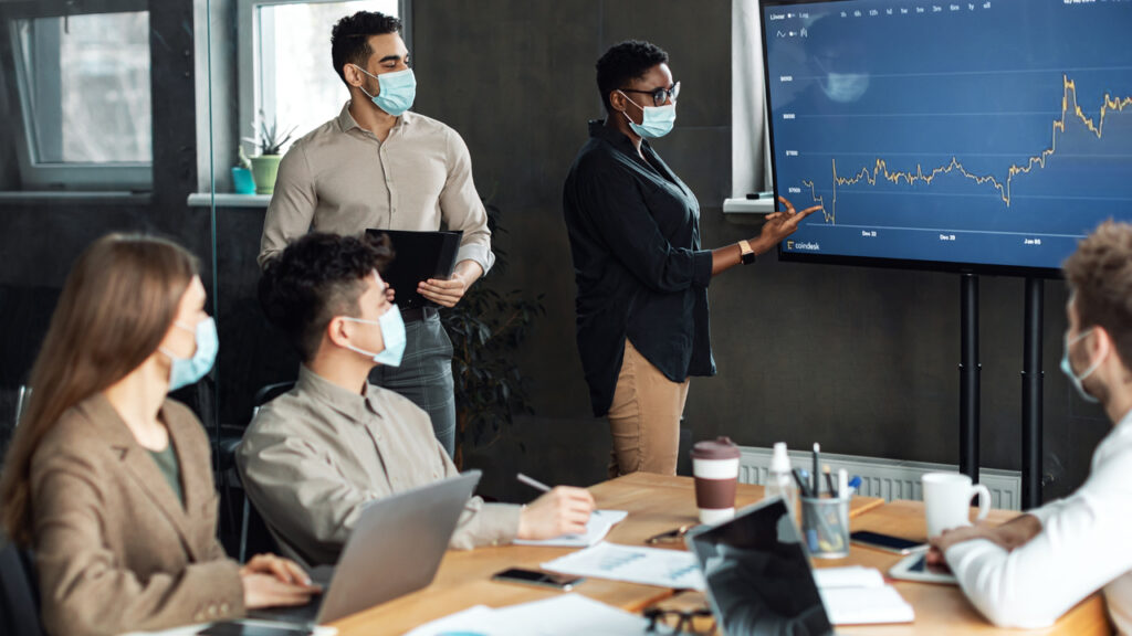 Briefing Concept. Black Businesswoman In Medical Mask Giving Speech During Seminar With Teammates In Office, Showing Financial Performance On Tv Screen Monitor Standing With Secretary In Board Room